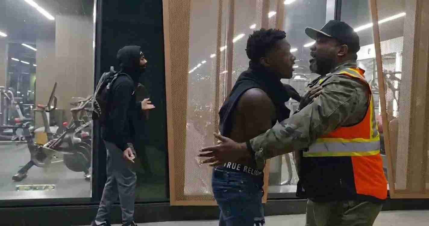 Security guard in a reflective orange vest holds back a shirtless man in a gym lobby as another person in a hoodie watches from the doorway.