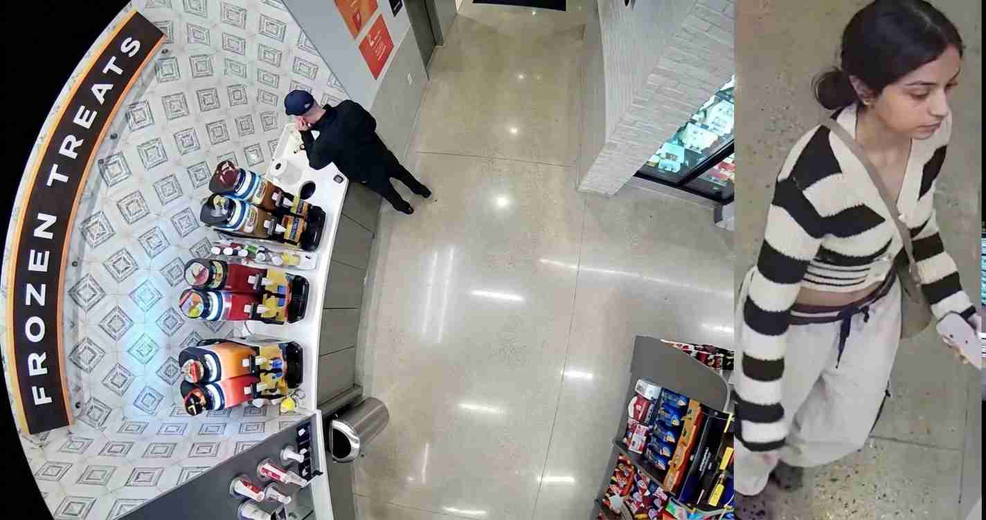 Man in dark clothing near a beverage display in a store; a woman in a striped sweater walks past on the right.