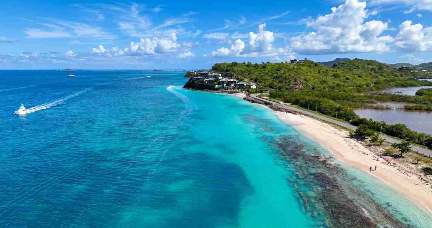 Aerial view of a tropical coastline with turquoise water, white-sand beach, and green hills with houses along the shore.