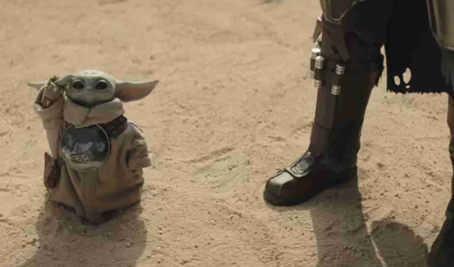 Grogu (Baby Yoda) in a tan robe stands in a dusty desert beside a booted armored figure.