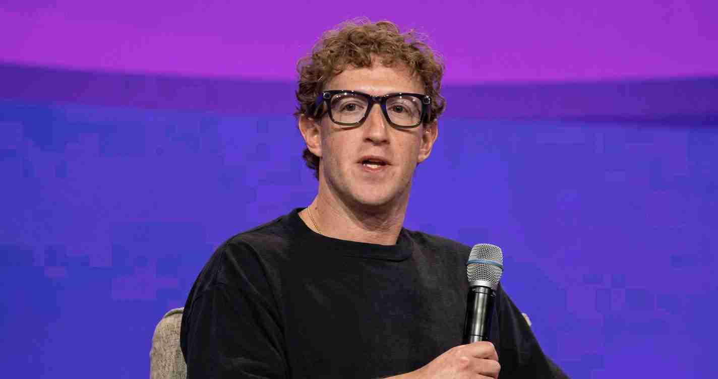 Man in a black shirt with glasses holds a microphone while speaking on a stage with a purple backdrop.