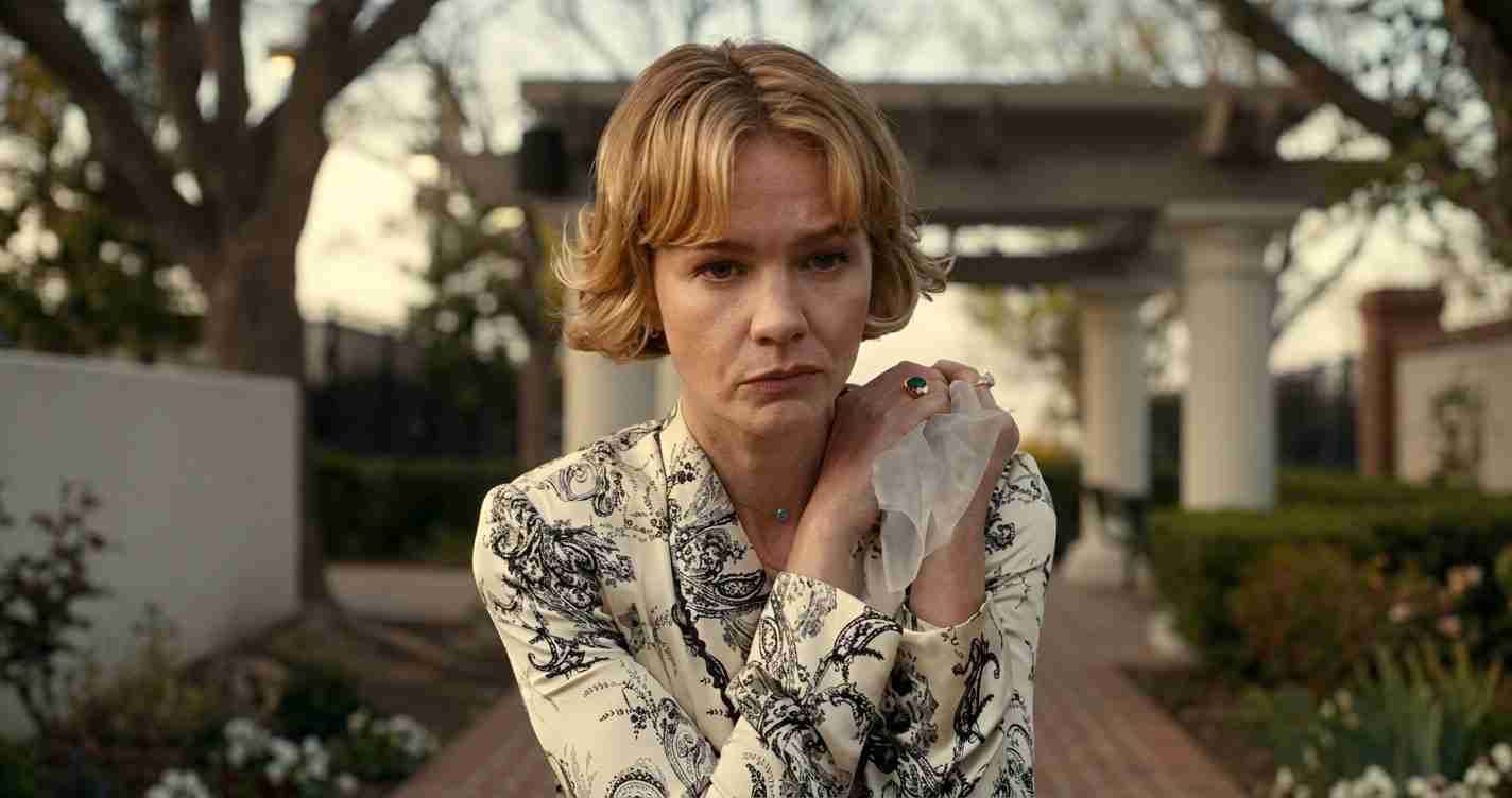 Woman with short blonde hair in a patterned blouse stands outdoors, looking pensive while holding a tissue near her chest.