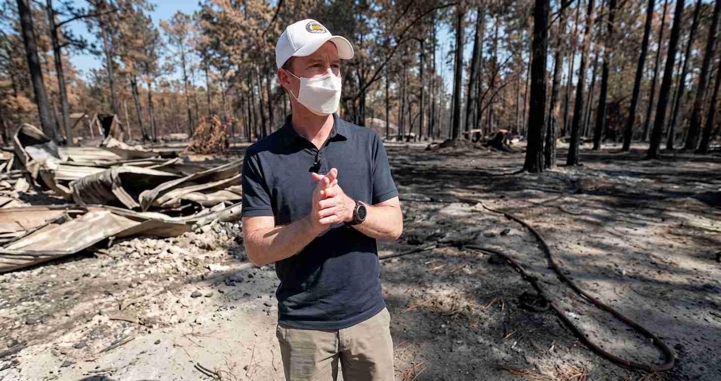 Man wearing a white cap and face mask stands in a scorched forest with charred trees and debris after a wildfire, hands clasped.