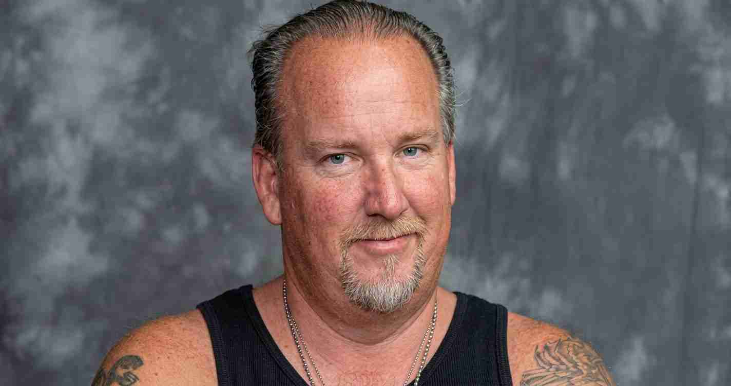 Portrait of a middle-aged man with a goatee, wearing a black sleeveless shirt, tattoos on both arms, against a gray backdrop in a studio portrait.