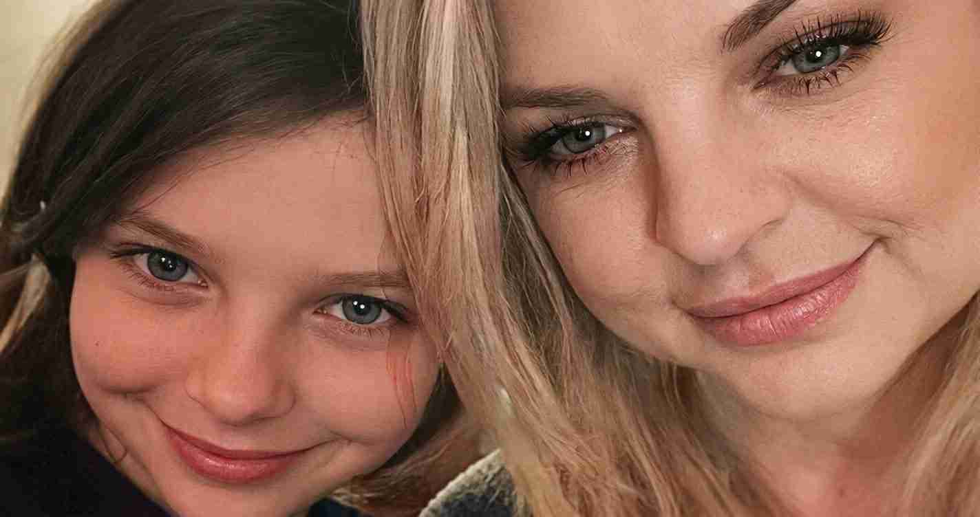 Close-up selfie of a smiling adult woman and a young girl sharing a moment, both looking at the camera.
