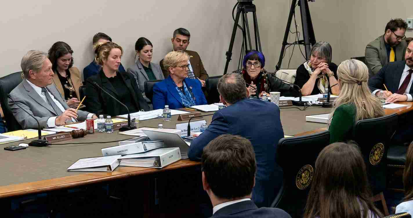 Formal committee meeting around a large table with microphones, documents, and nameplates in front of each participant.