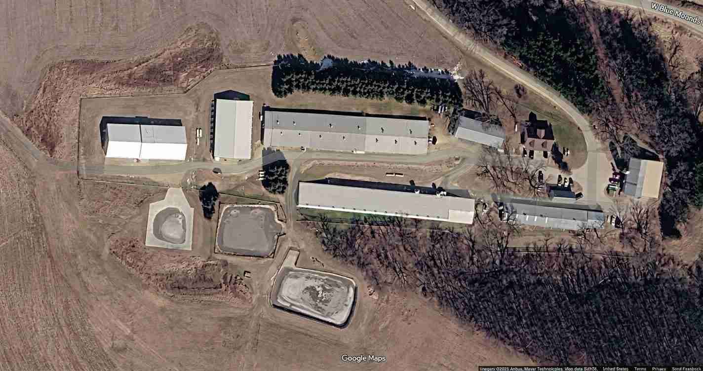 Aerial view of an industrial complex with several long beige buildings, circular concrete pads, and surrounding trees on open land.","Satellite image of a facility lot with multiple rectangular buildings and a curved road network.","Overhead view of a campus-like industrial site with long warehouses, open lots, and treeline nearby.","Top-down view showing a cluster of long rectangular buildings and circular pads in a rural setting.","Google Maps satellite image of a small industrial park with linked warehouses and yard areas.