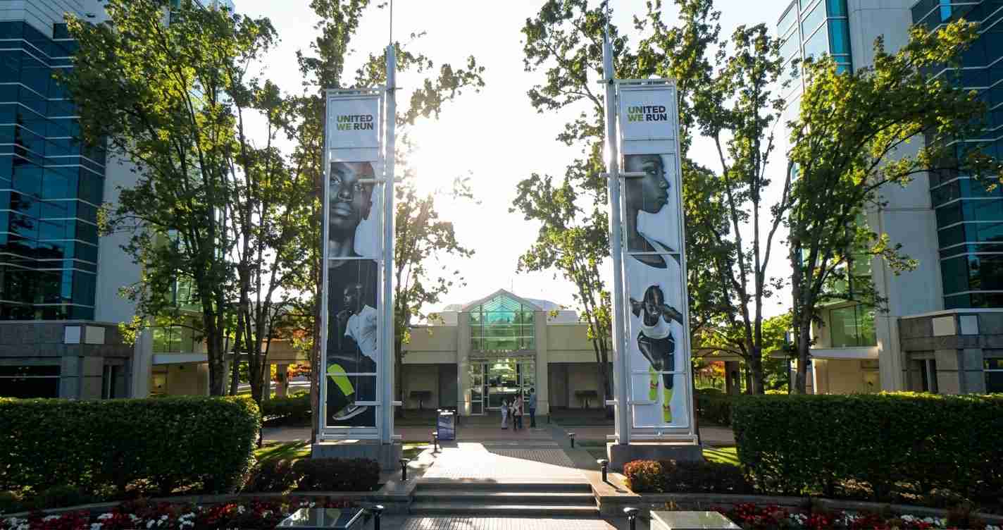 Entrance to a modern campus building with tall banners reading 'United We Run' flanking a glass atrium, trees and landscaping visible in foreground.