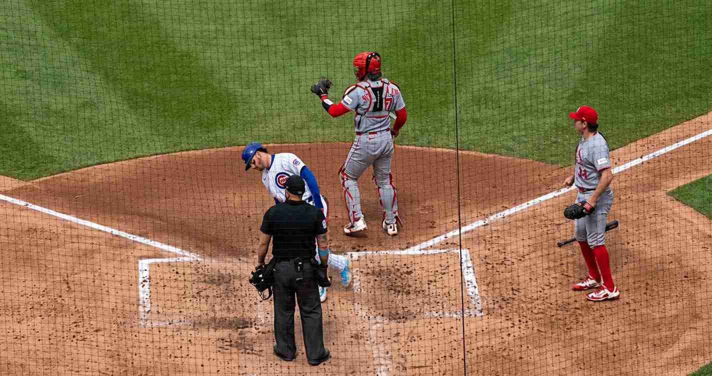 Baseball scene at home plate: umpire, catcher, and two runners near the plate with umpires in black, netting over the field.