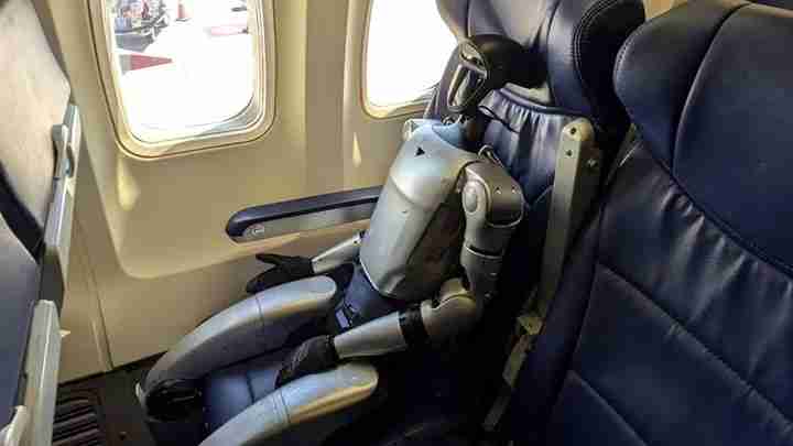 A silver humanoid robot sitting in an airplane seat by the window, showing articulated metallic limbs and a bulky torso.