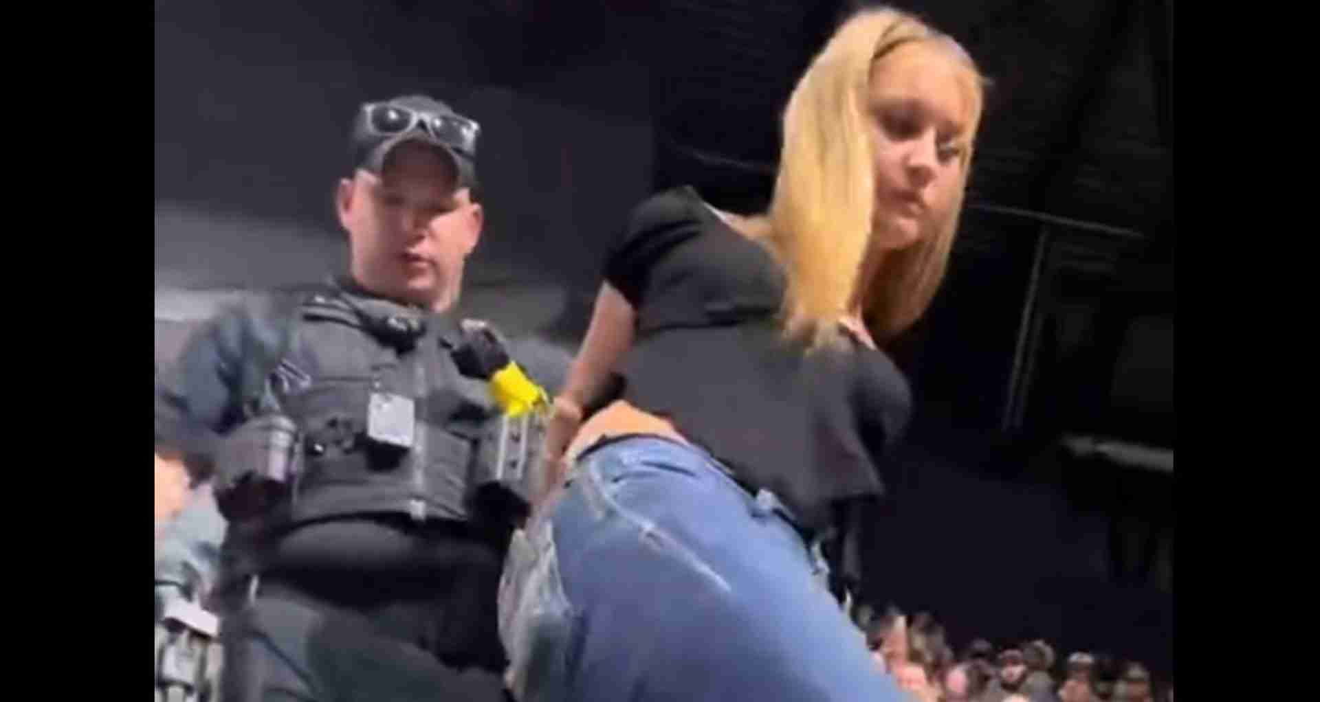 Security guard in a vest and cap stands on a dim stage near a crowd at a concert. a blonde woman in a black top leans toward the audience in the foreground.