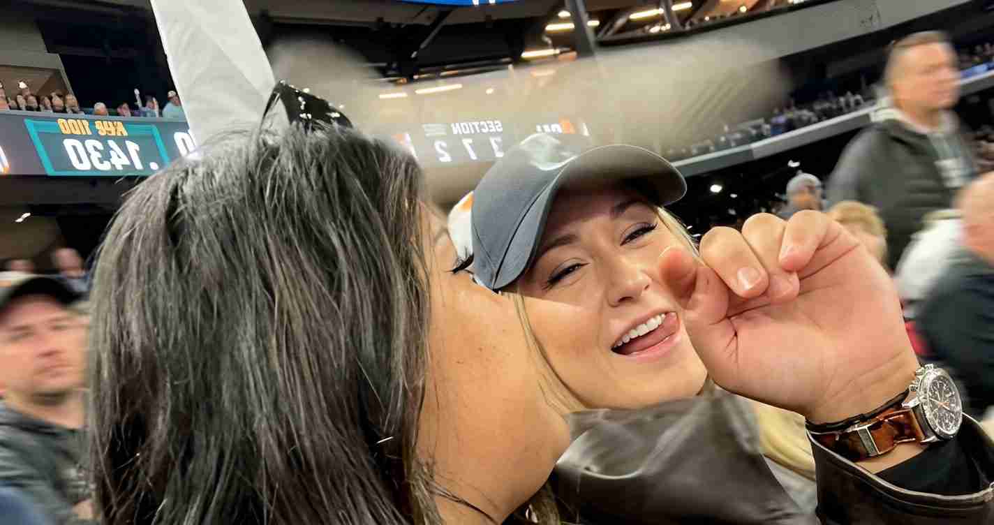 Two people at a crowded sports arena share a quick kiss on the cheek as they smile at the camera, one wearing a gray cap and a watch nearby.