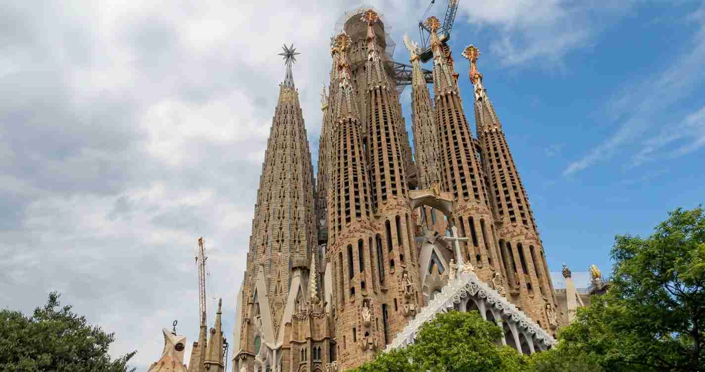 Sagrada Família’s tall stone towers under a partly cloudy sky, with ornate Gothic details and green trees in the foreground