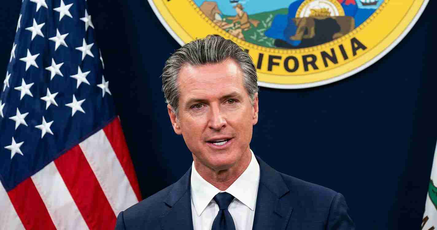Male politician in a dark suit and tie speaking at a press briefing, with the California state seal and an American flag in the background.