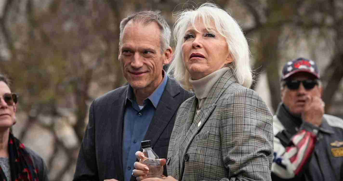 Woman with white hair wearing a gray plaid blazer holds a bottle while standing beside a man in a dark suit outdoors, with others in the background.