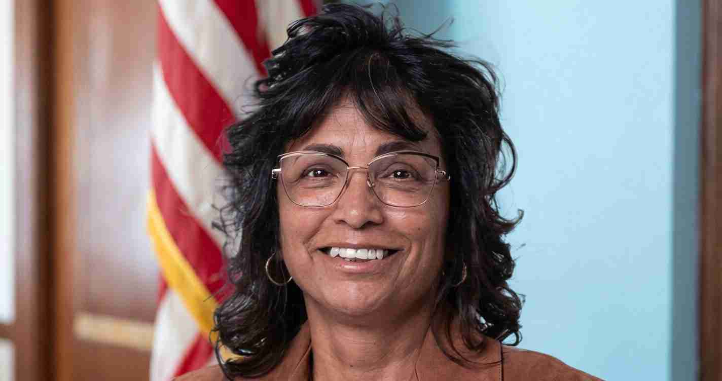 Portrait of a smiling woman with short dark hair and glasses, indoors with an American flag in the background.
