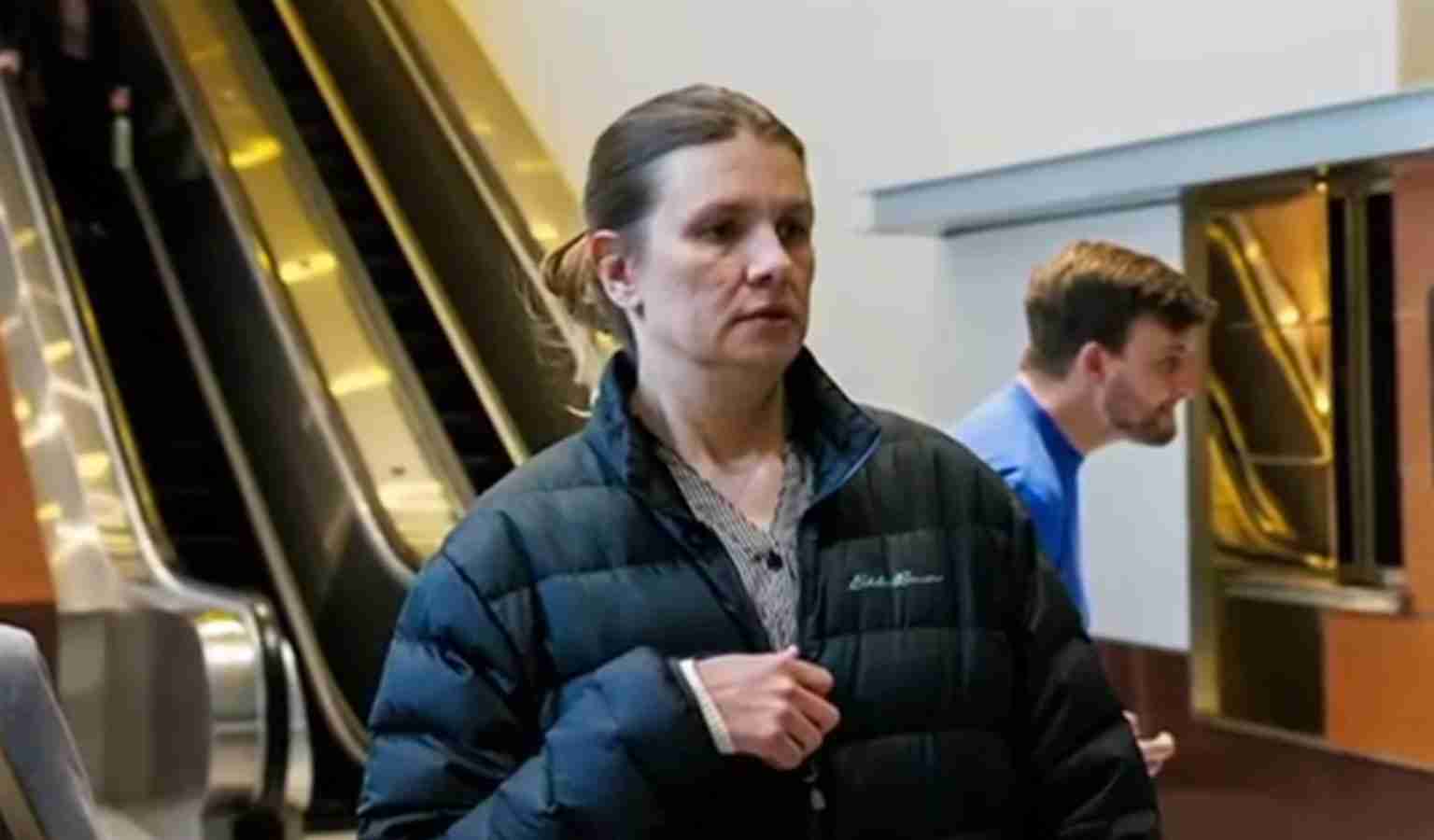 Woman in a dark puffer jacket standing near an escalator in a mall or transit hall, looking ahead with a neutral expression.