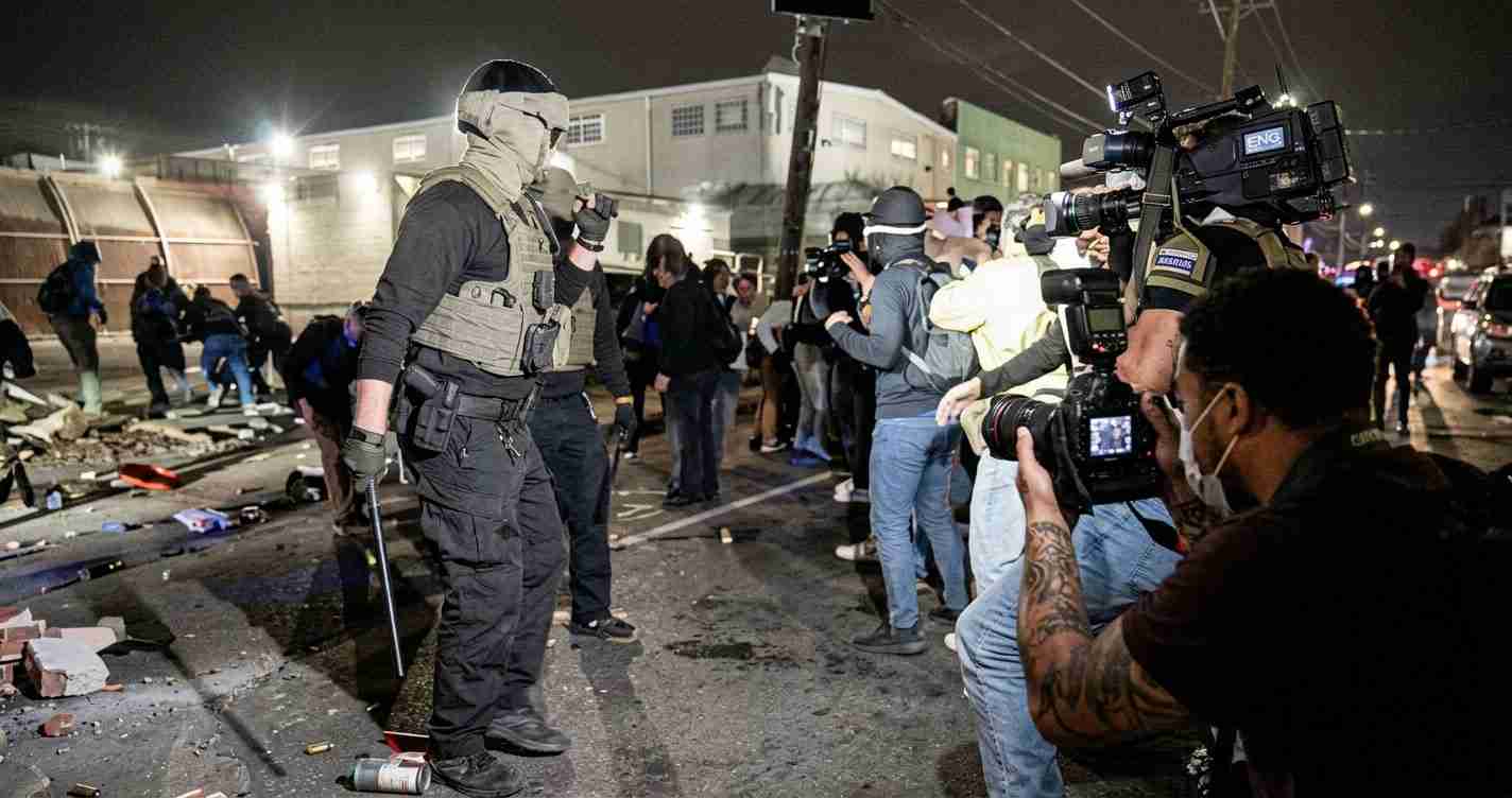 Armed officer in tactical gear stands on a debris-strewn urban street at night as journalists film the scene with large cameras nearby.
