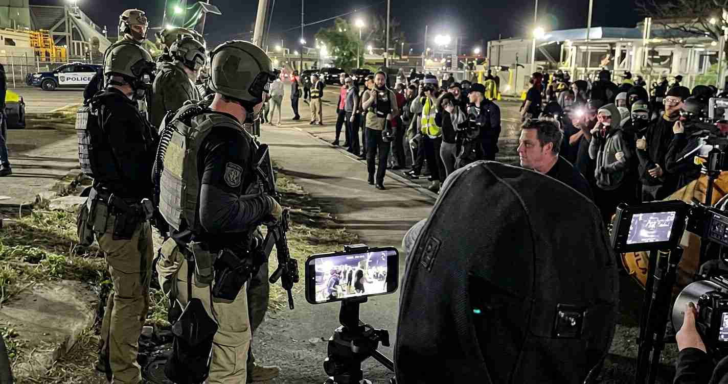Nighttime scene of heavily armed police in tactical gear facing a crowd of protesters and reporters; cameras and phones point toward the officers.