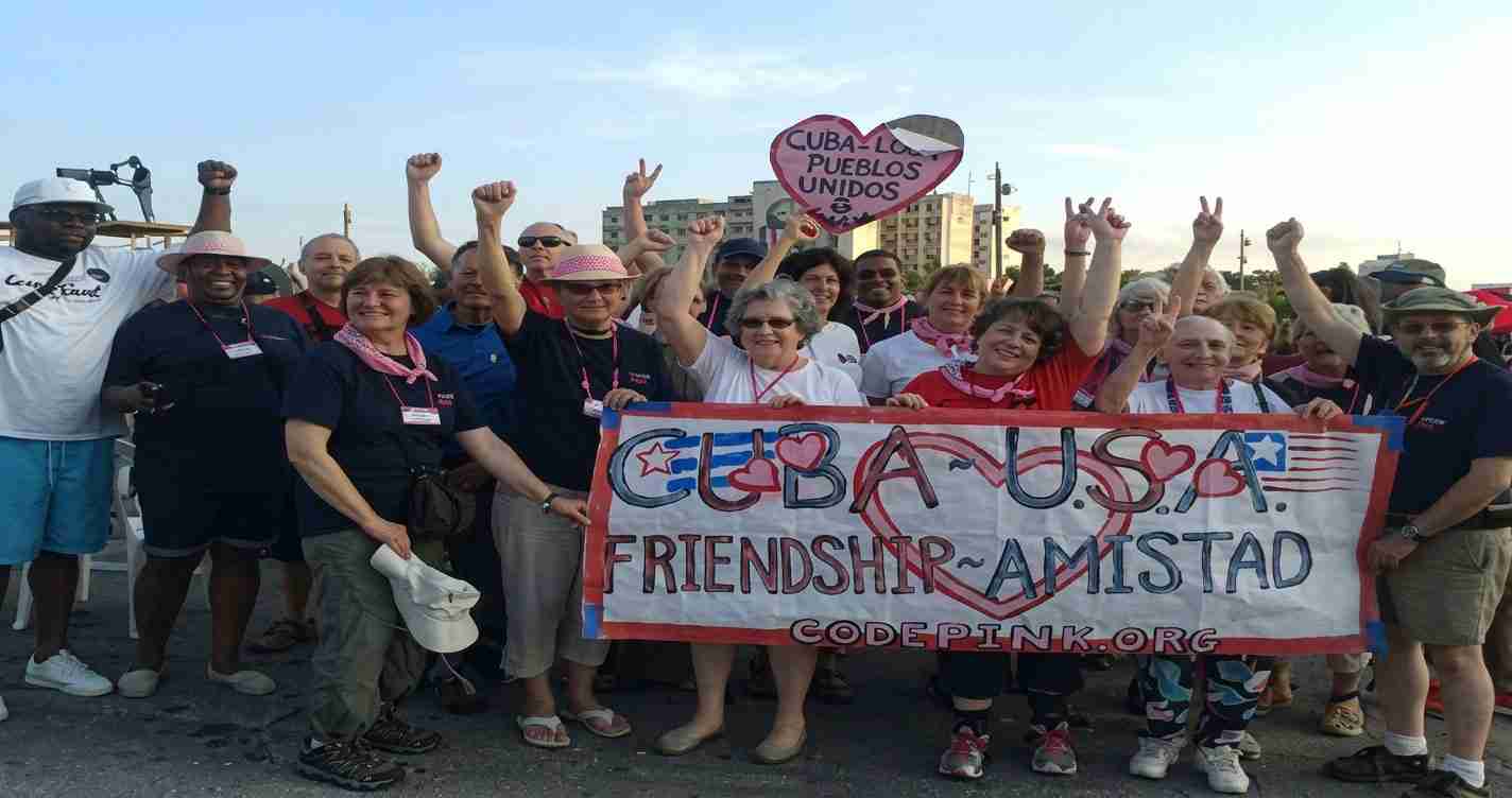 Group of diverse people at a unity rally holding a banner reading 'CUBA USA FRIENDSHIP AMISTAD' with hearts and US/Cuba symbols in the background.