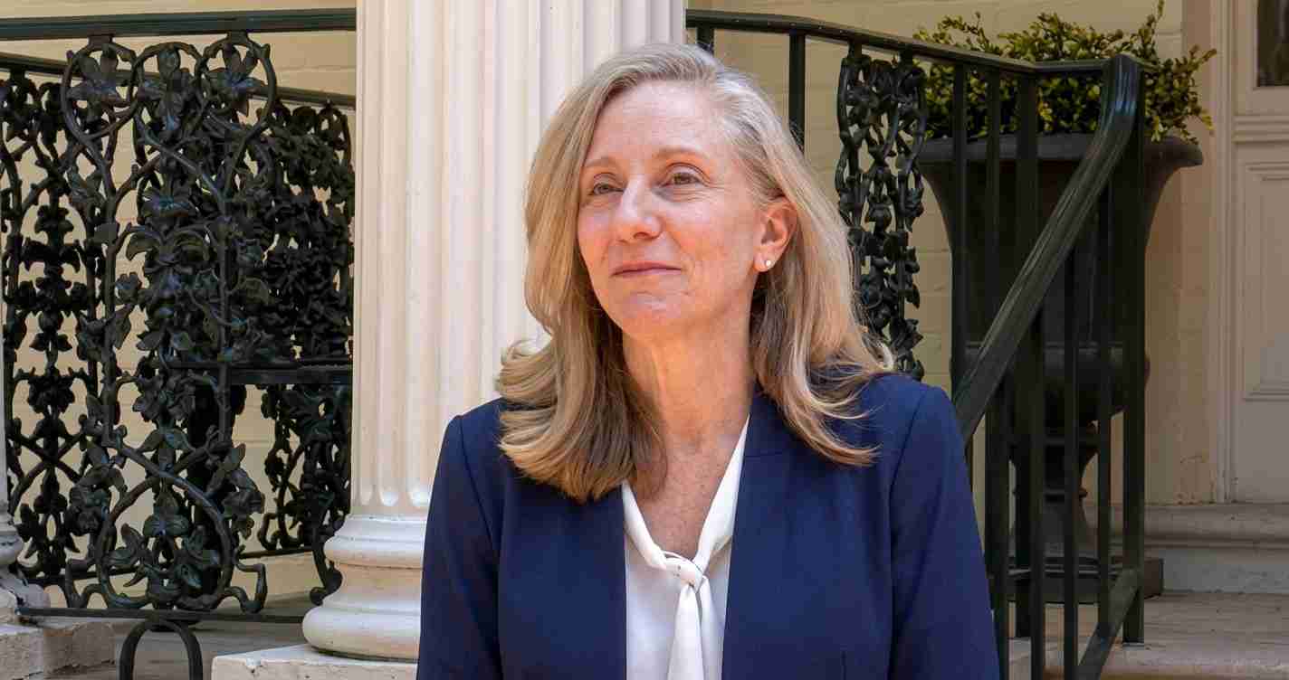 Professional woman with shoulder-length blonde hair in a navy blazer and white blouse, seated on steps outside a building.