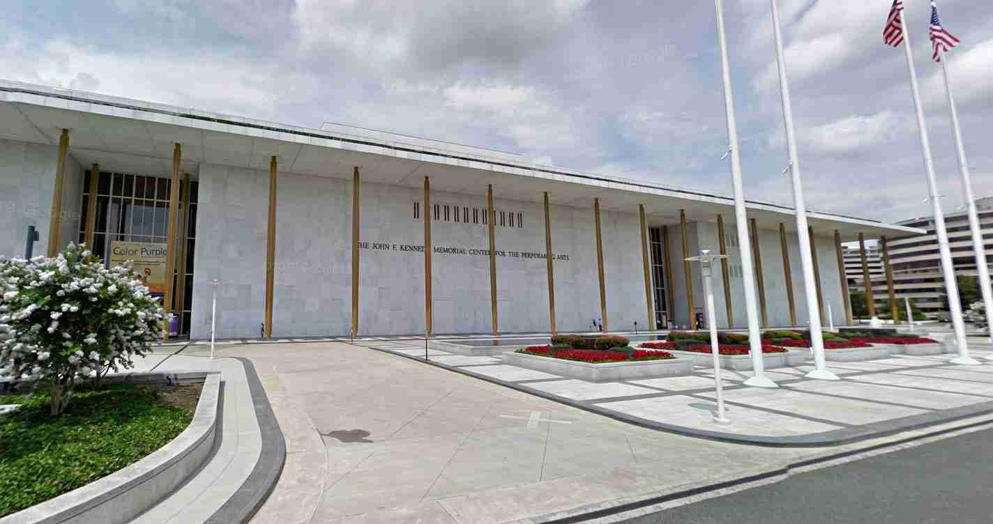 Front view of a memorial center with tall white columns, flagpoles, and a landscaped plaza with red flowers.