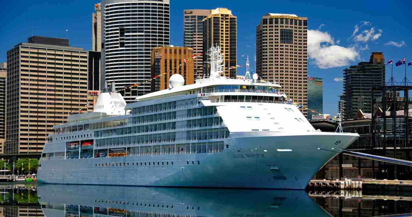 Large white cruise ship moored at a dock with tall city buildings in the background.