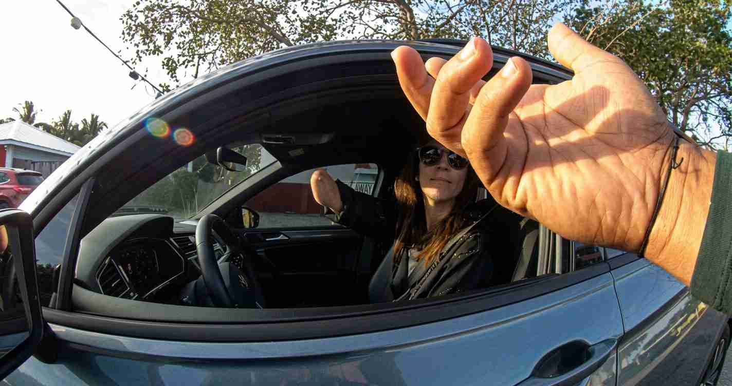 Person in a blue car reaches an arm toward the camera, taking a selfie from the driver’s seat with sunglasses on in the background and lens flare at the left edge.