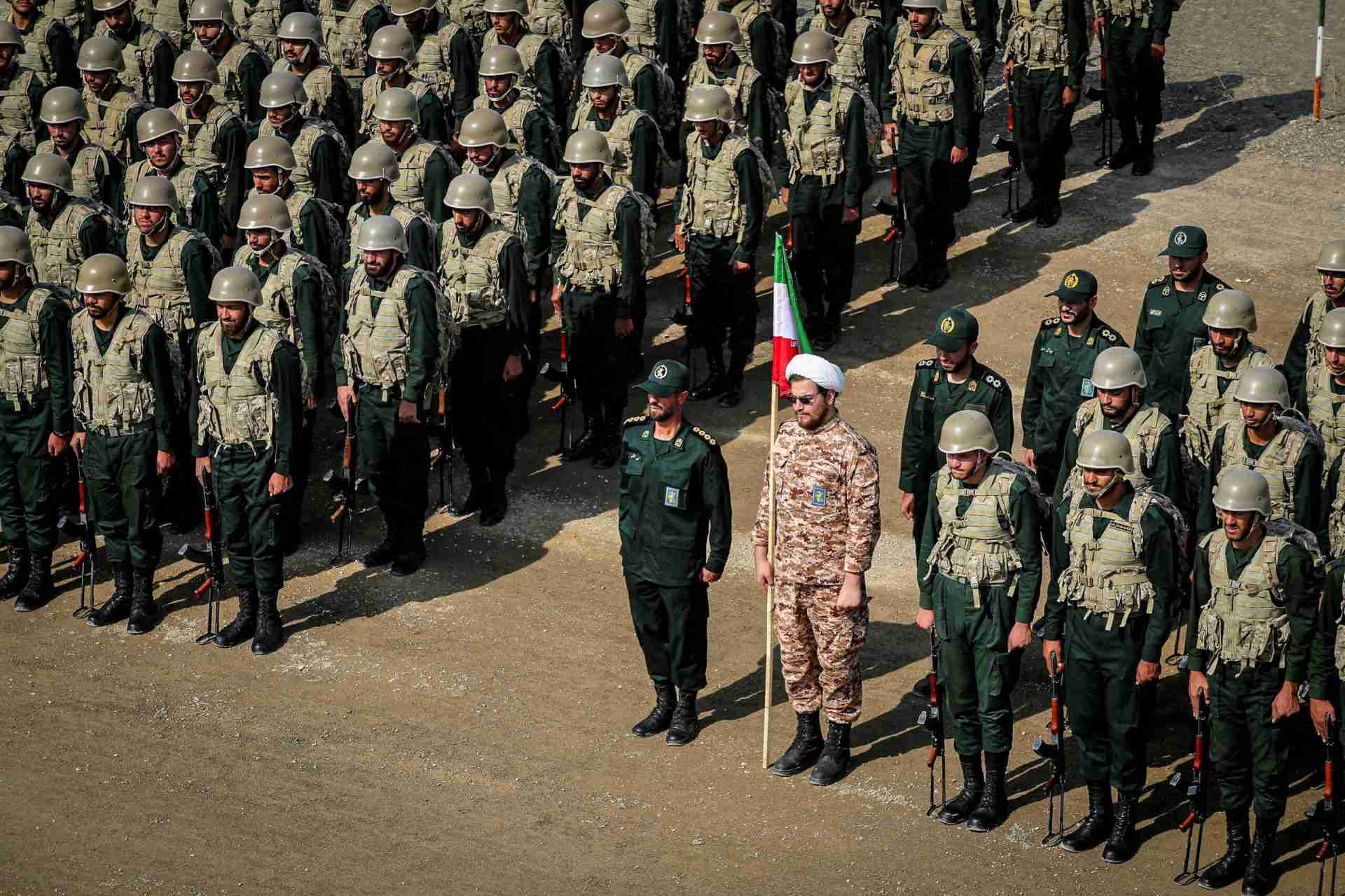 Rows of soldiers in green camouflage uniforms and helmets standing in formation outdoors, a flag bearer in front.