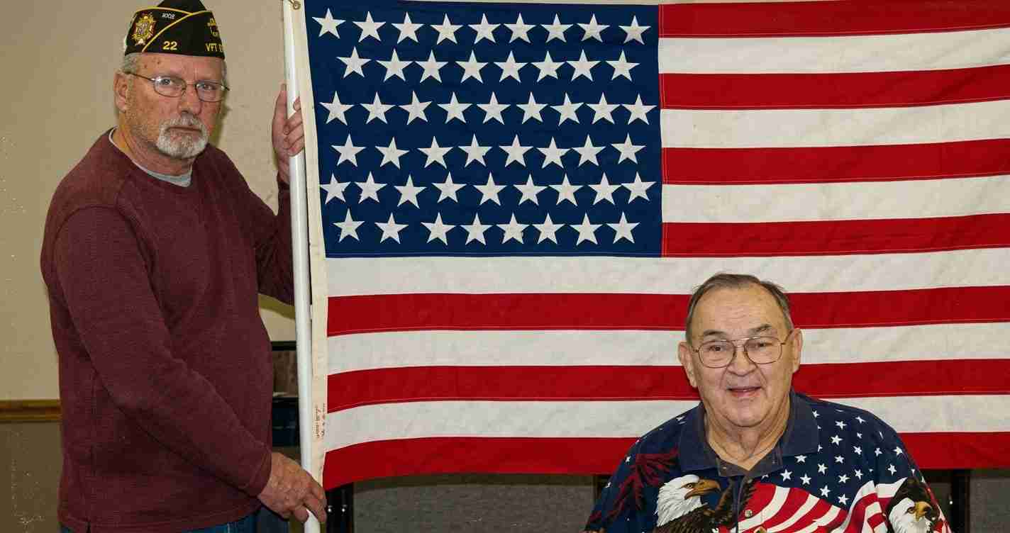 Two older men pose beside a large American flag; one man stands left wearing a veterans cap and maroon sweater, the other sits right in a patriotic shirt.
