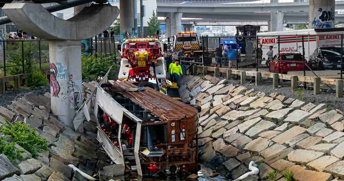 Overturned brown bus wedged in a rocky canal after a crash, with rescue workers and emergency vehicles under an elevated highway.