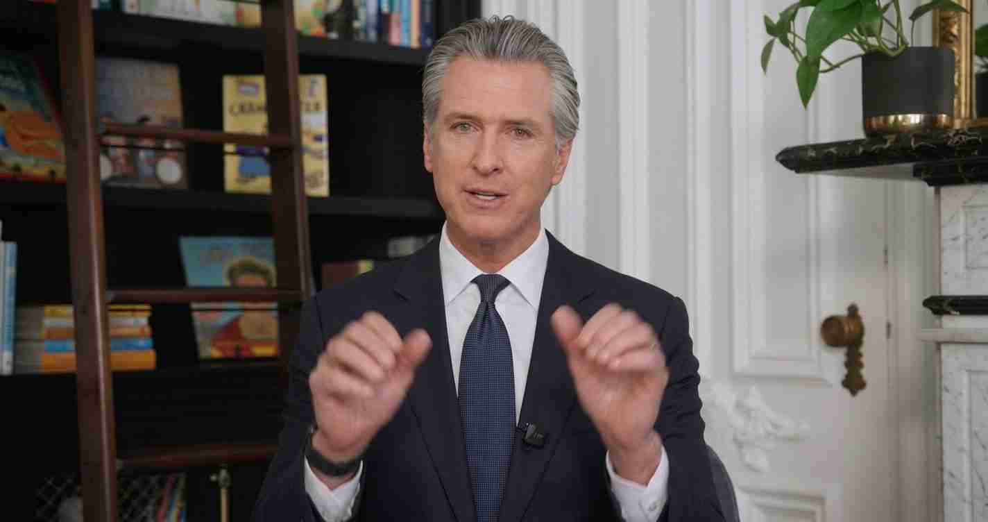 Well-dressed man in a dark suit speaks to camera, hands raised in a gesture, with a bookshelf behind him.
