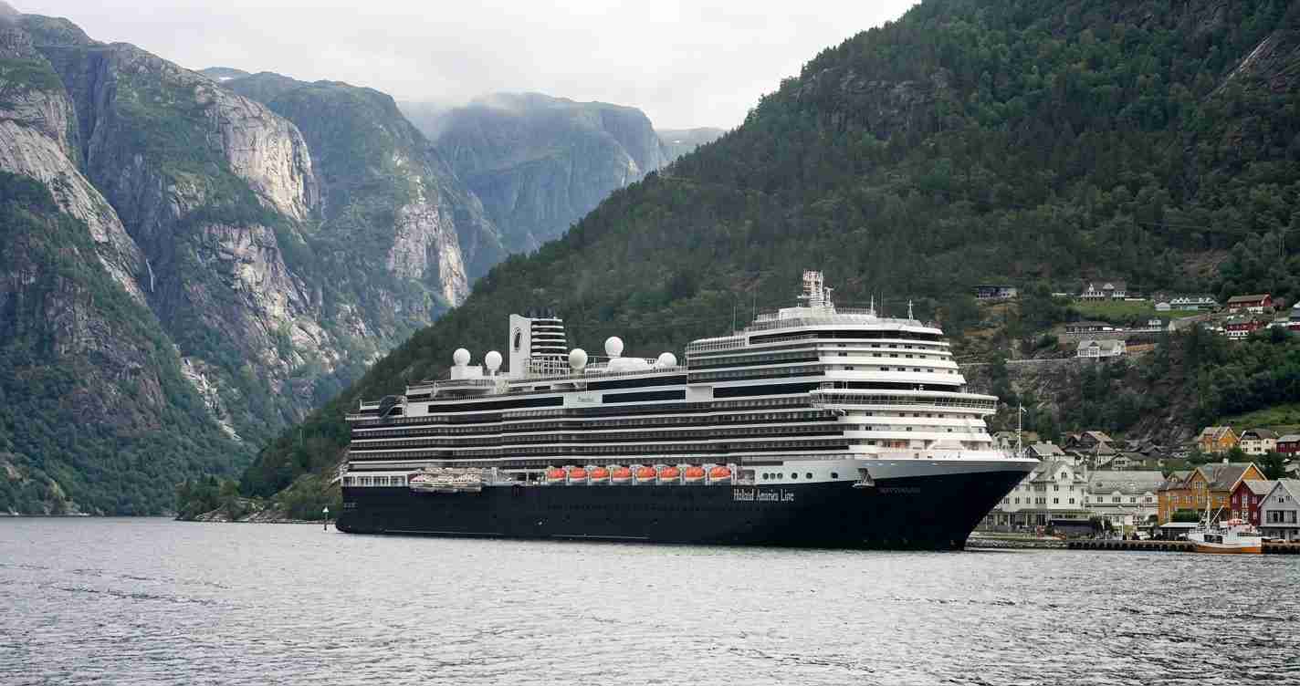 Holland America Line cruise ship anchored in a narrow fjord, with steep green mountains and a small town along the shoreline behind it.