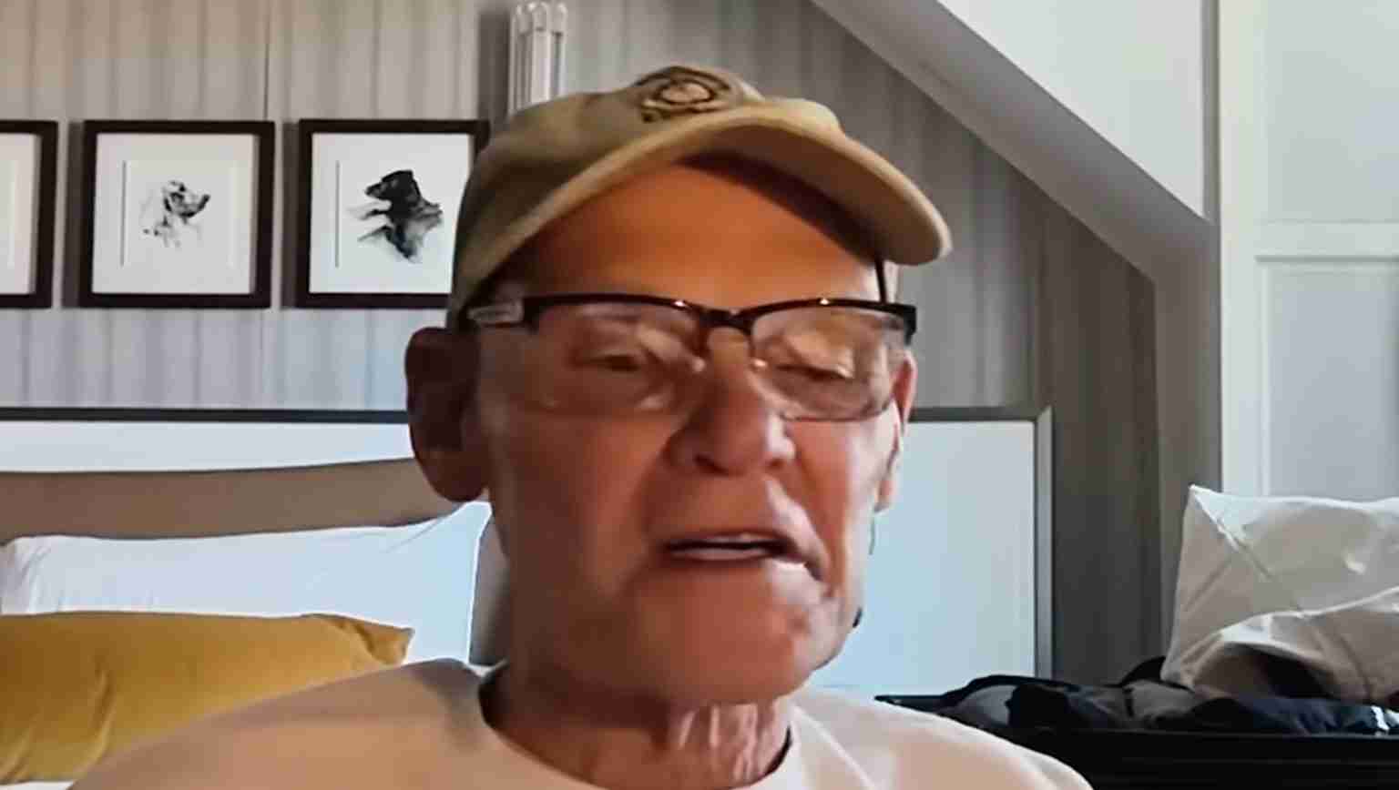 Older man wearing glasses and a beige cap, speaking during a video call, with framed dog pictures on the wall behind him.