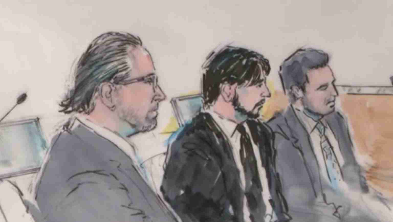 Three men in suits seated at a courtroom table, one with glasses on the left, others in the middle and right.