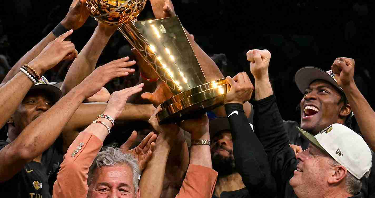 Basketball players celebrate, lifting a large gold trophy above their heads.