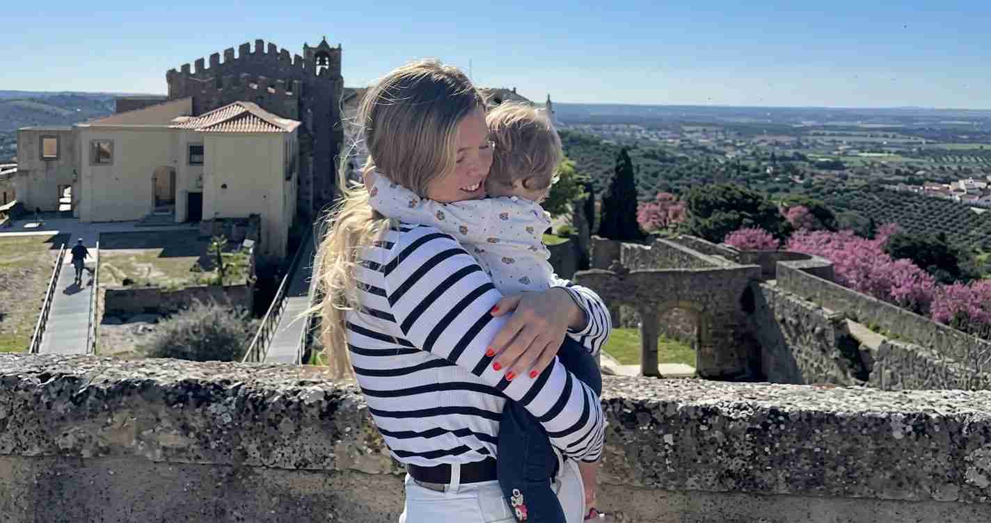 Woman in a striped shirt hugs a child on a stone wall with a medieval fortress and countryside in the background.