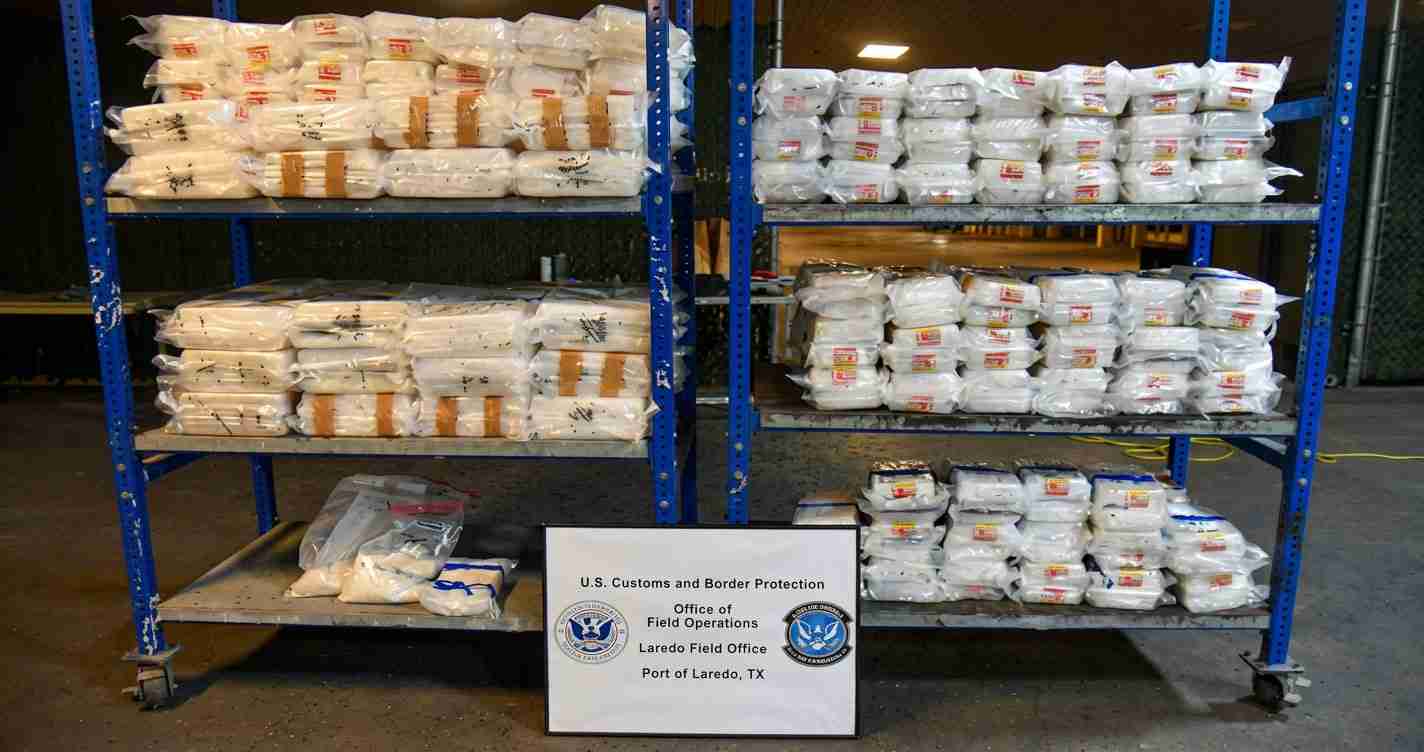 Warehouse scene with blue metal shelves stacked with white bags and a sign in front from U.S. Customs and Border Protection, Port of Laredo, TX
