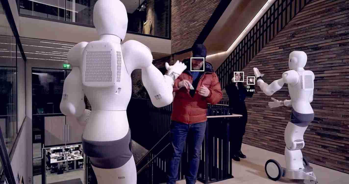 A man in a red jacket high-fives two white humanoid robots in a modern lobby with a wooden wall backdrop.