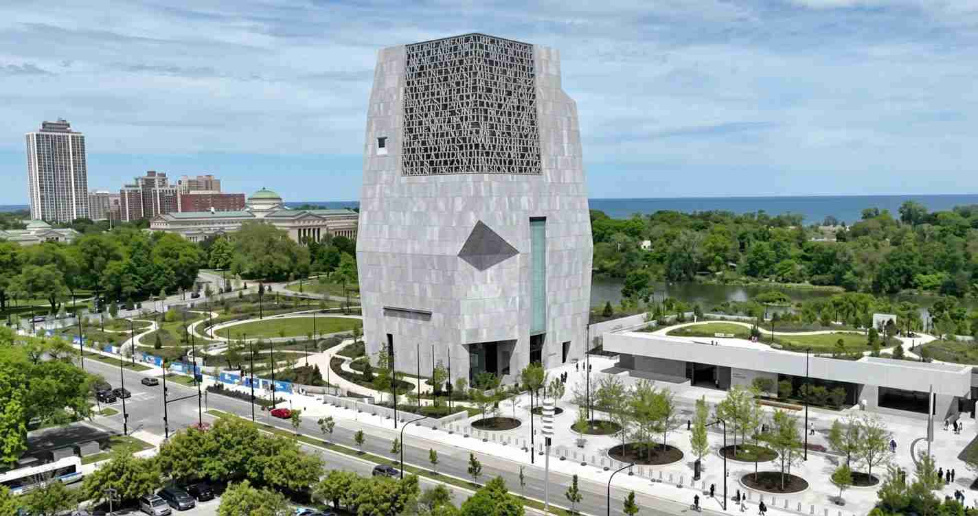 Tall angular gray stone tower rises from a landscaped park with winding paths, near a waterfront.