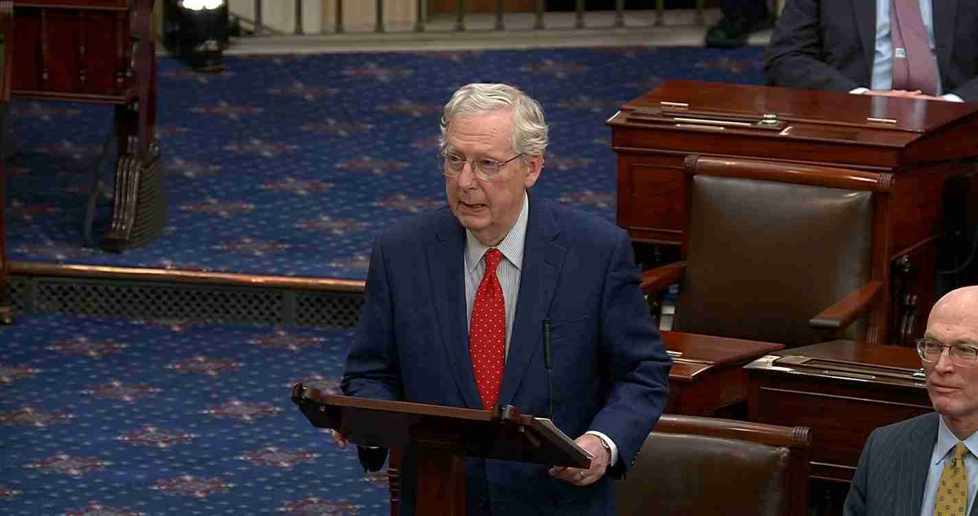 Elderly man in a navy suit and red tie speaks at a wooden podium in a legislative chamber; blue carpeted floor and desks around him.
