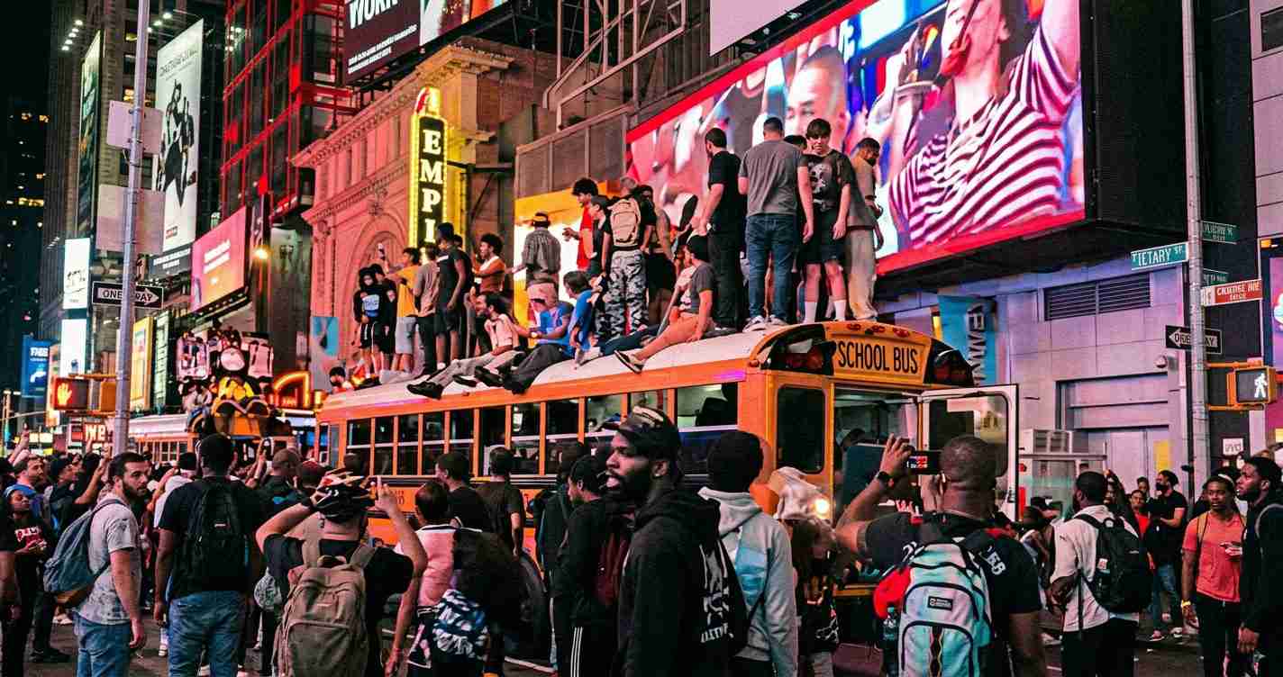Crowd gathered on an orange school bus in Times Square at night, with towering neon billboards and pedestrians around.