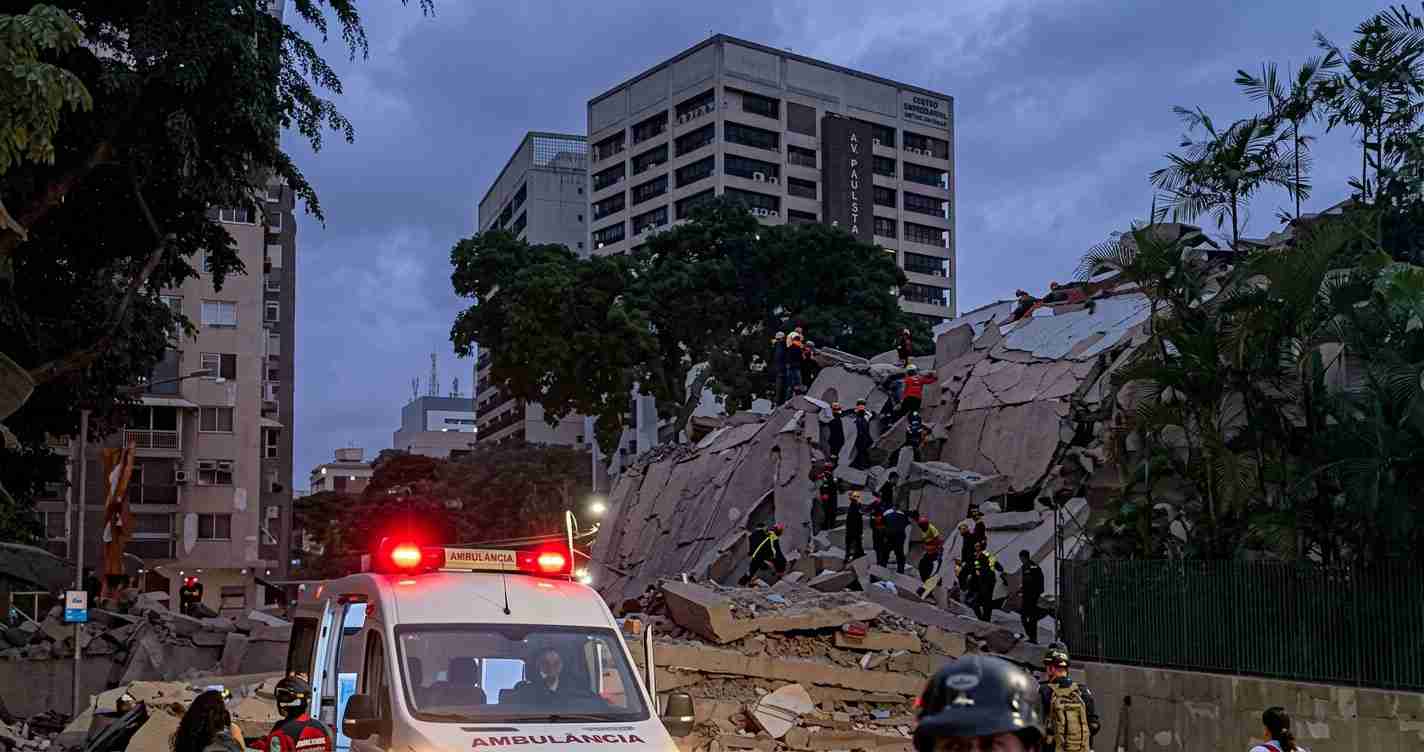 Emergency responders scan a collapsed concrete structure at dusk, with an ambulance in the foreground and rescuers on rubble.