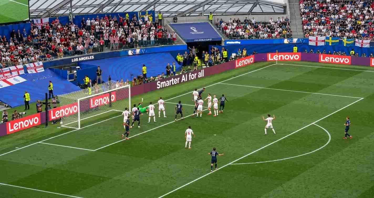Soccer match near the goal: white and navy teams contest a set-piece as players cluster by the net with a goalkeeper in green. Crowd watches from stands; Lenovo ads line the pitch edge.