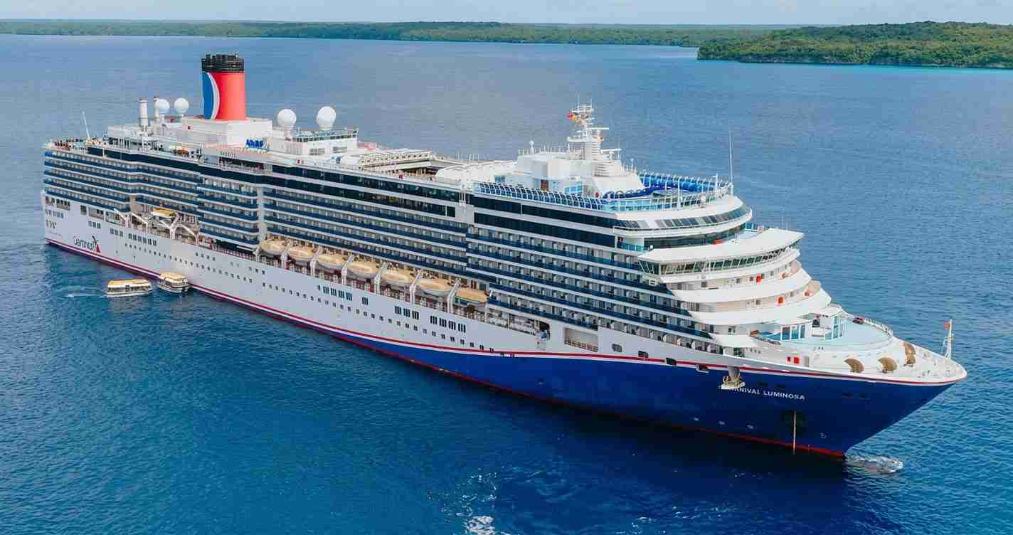 A large white and blue cruise ship with multiple decks sailing in calm blue water, red funnel visible at the top.