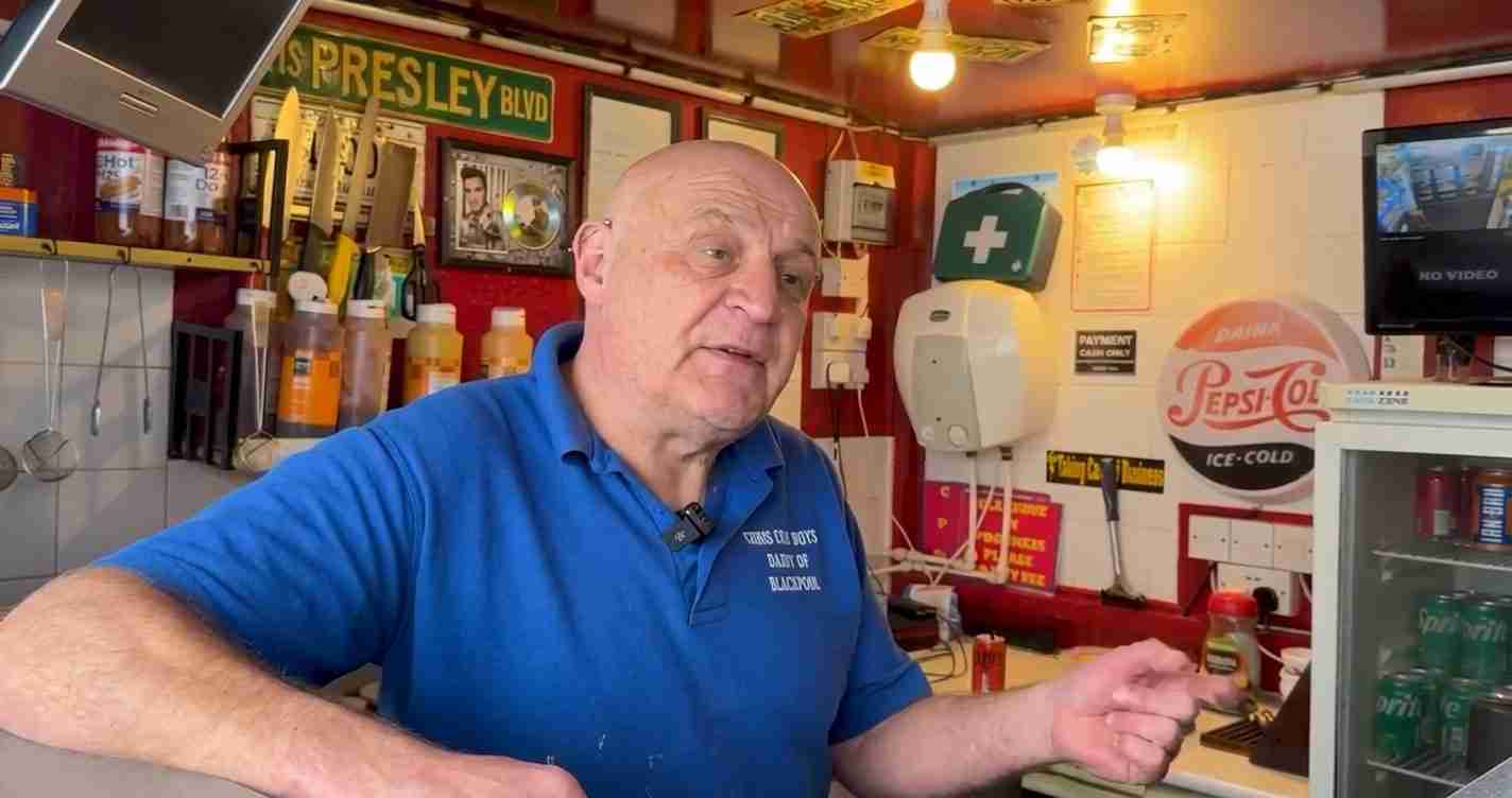 Bald man in a blue polo behind a counter in a retro shop with bottles and vintage signs.