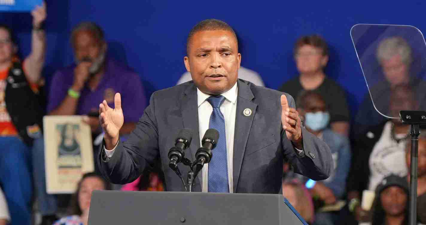 Man in a suit speaks at a podium with two microphones, gesturing with both hands in front of a blue backdrop.