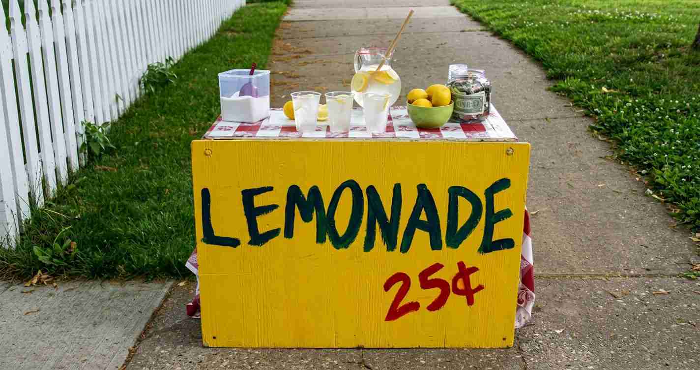 Street-side lemonade stand with a bright yellow sign reading 'LEMONADE 25¢', cups, pitcher, lemons, and a jar of coins on a checkered tablecloth on a sidewalk.
