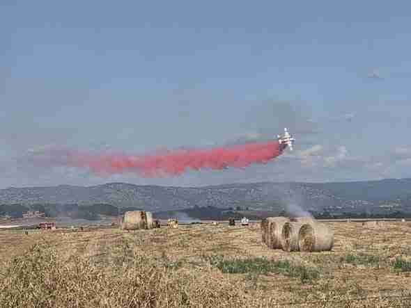 Small airplane trailing red smoke flying low over a rural field with hay bales and distant hills in the background.