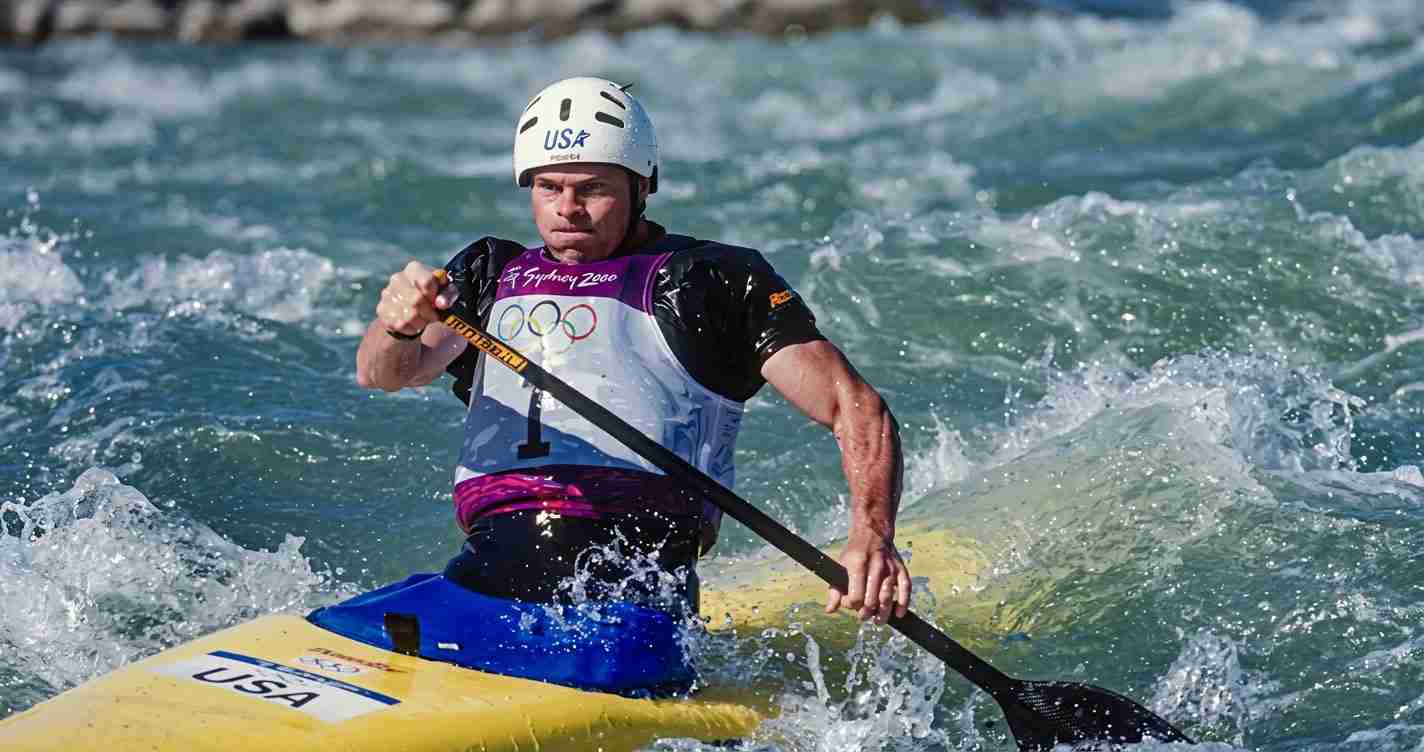 Male kayaker wearing a white USA helmet and bib paddles a yellow kayak through rough whitewater waves with a double-blade paddle within reach of the camera.